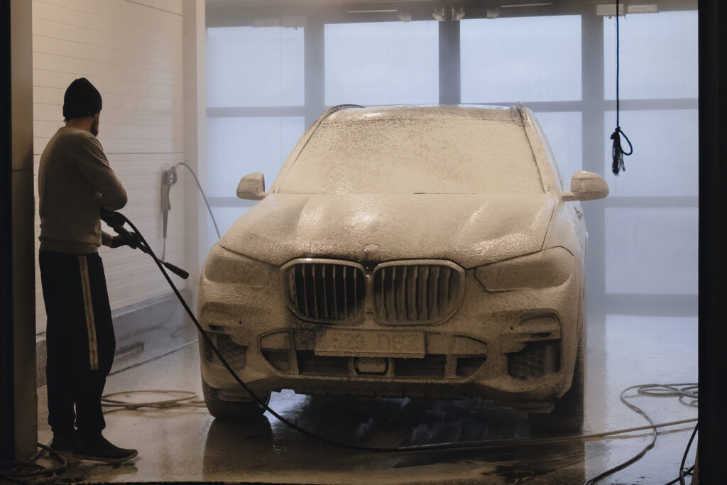 BMW X5 kaetud aktiivvahuga käsipesu boksis – töötaja loputab sõidukit survepesuriga enne detailset puhastust.